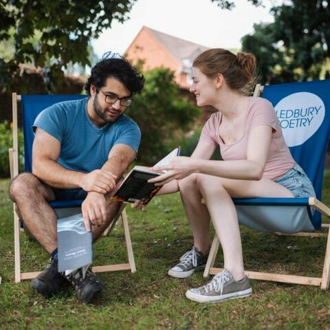 A male and female sitting on Ledbury Poetry branded deckchairs and reading a book together