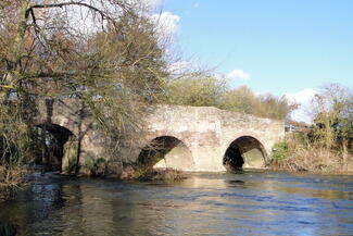 ancient bridge with river flowing through, trees to one side and bright blue skies