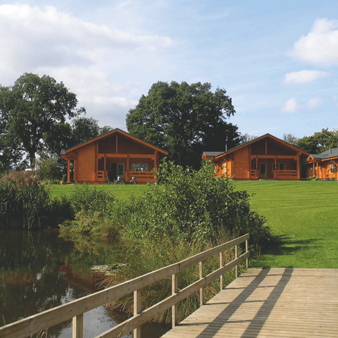 view across wooden footbridge looking towards log cabins in green field surrounded by trees