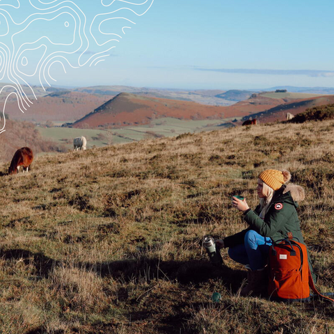 Woman sat on hilltop with grazing ponies in the distance