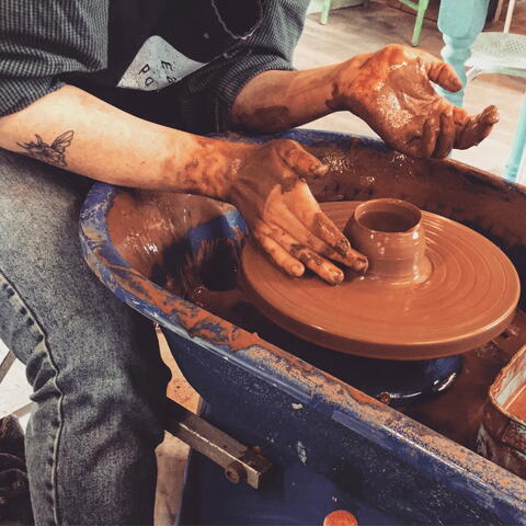 person sat with clay-covered apron making red clay pot on potters wheel