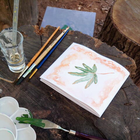green leaf painting on white paper pad, with paint brushes, glass of water on a outdoor wooden table with wooden tree stump stool