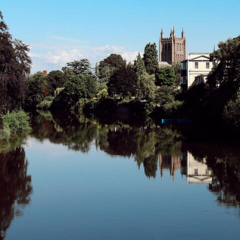 A view across a very still River Wye towards the Cathedral with the building, blue skies and trees reflected in the water